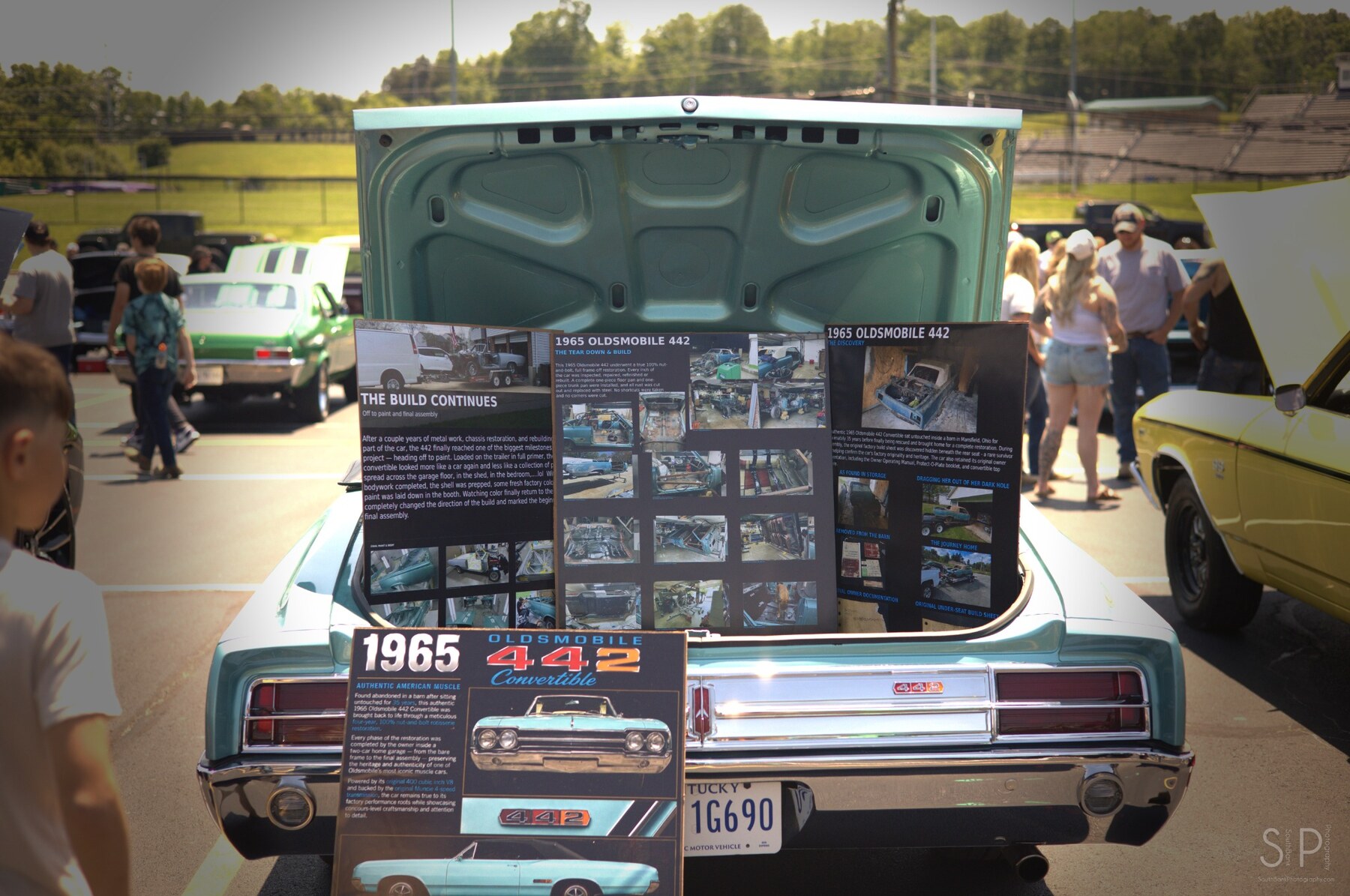 Rear of a turquoise 1965 Oldsmobile 442 with a restoration display in the open trunk